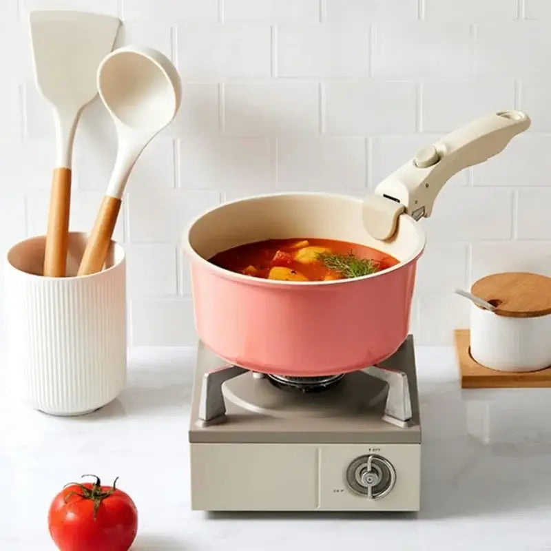 Casserole rose avec Poignée Amovible Universelle sur un réchaud, accompagnée de spatules en cuisine