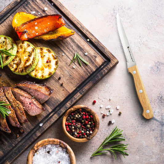 Set 4 à 8 Couteaux à Steak Dentelés – Manche Bois Naturel posé près de viande et légumes grillés sur planche en bois